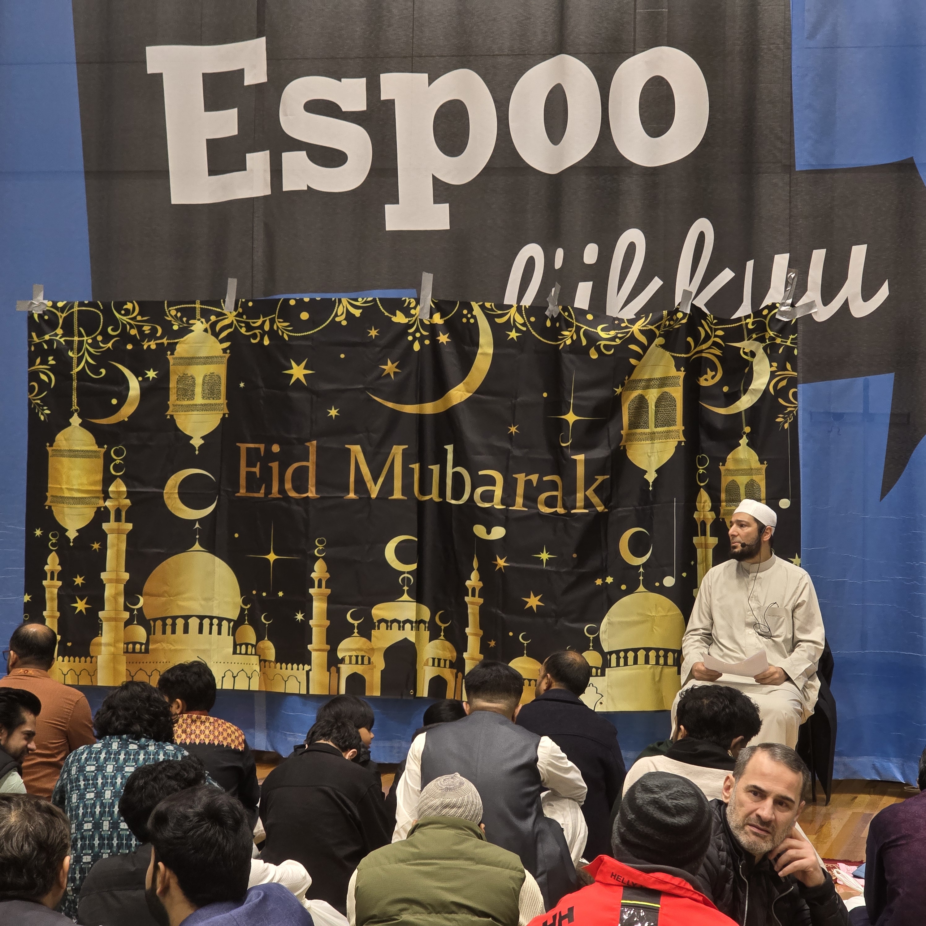 A speaker addressing seated attendees in front of an 'Eid Mubarak' banner at an EMC gathering